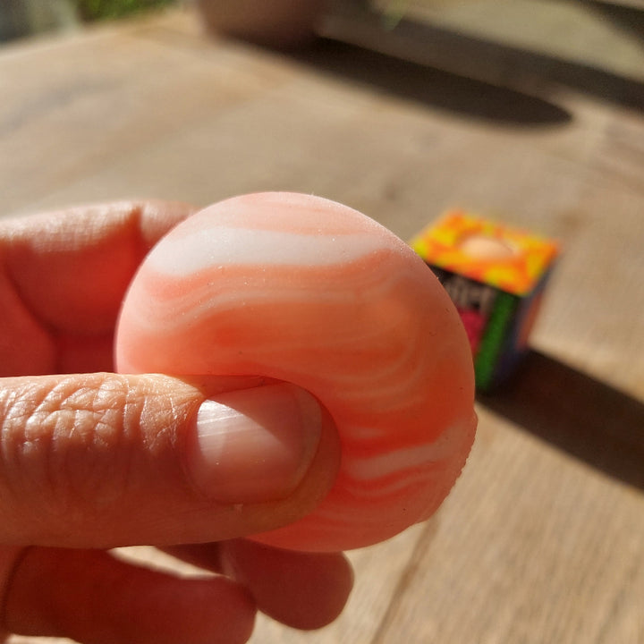 oranje teenie needoh, kleine needoh in zon tussen duim en vinger, achtergrond doos van needoh teenie, motoriek fidget