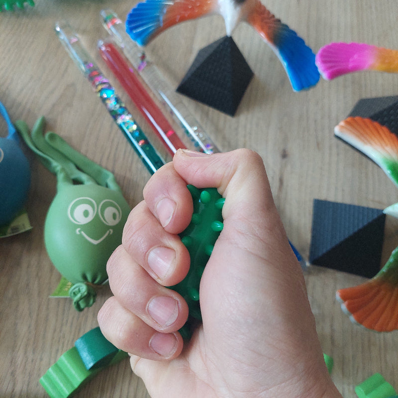 Hand knijpt in een massage-olijf met voelbare stekeltjes voor sensorische afleiding tijdens een medische behandeling, met de overige materialen uit de set op de achtergrond.