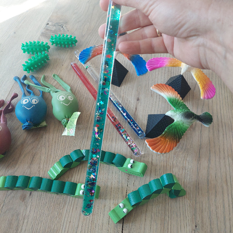 Hand houdt een toverstaf vast voor visuele afleiding tijdens vaccinatie of bloedprikken, met de complete afleidingsset op de achtergrond.