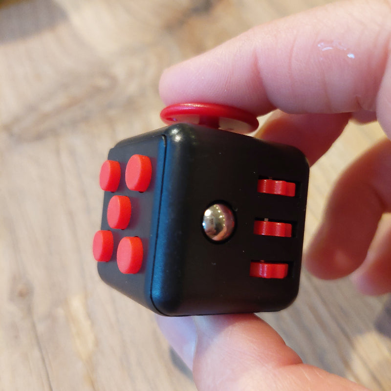 zwarte fidget cube met rode schakelaars, knoppen en draai , hand hout fidgetcube vast boven houten tafel, motoriekspeelgoed