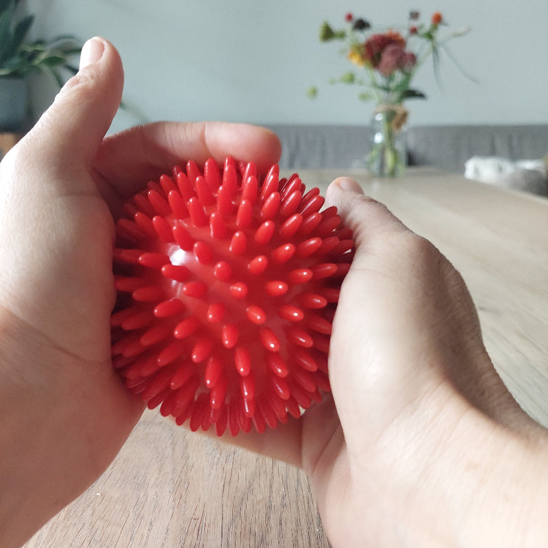 roter massageball in händen, an einem Holztisch mit blumen im hintergrond, sensorischer massage ball, 