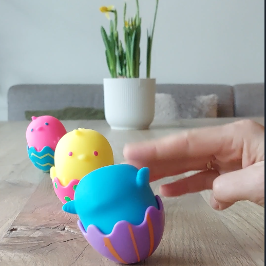 drie ChickaDeeDoos op houten tafel met paasbloemen op de achtergrond, twee kuiken kijken, een rolt op de kop met hand die duwt